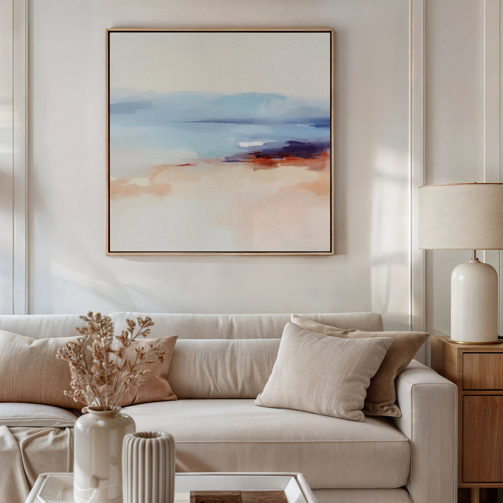 A modern living room with a beige sofa, light cushions, dried flowers in a vase, and a table lamp on a wooden side table, featuring the Windswept Coastline Canvas Art - Square in blue, peach, and cream tones for a tranquil ambiance.