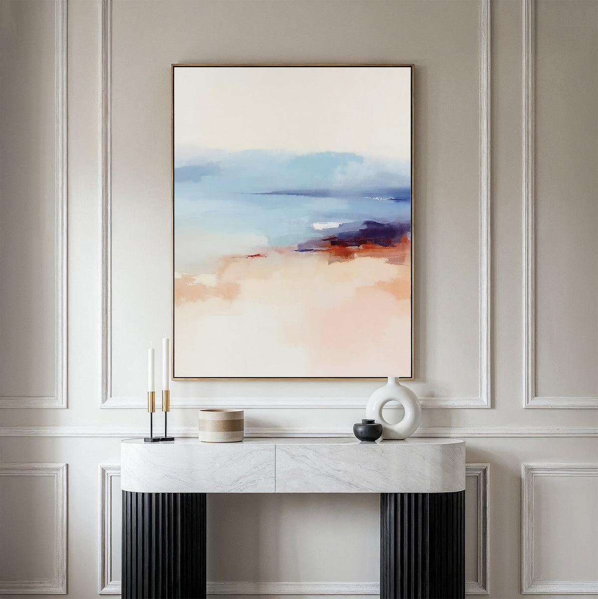 A minimalist room with a white paneled wall, a marble console table with decor, and the Windswept Coastline Canvas Art - Vertical in soft blue, beige, and coral tones above, creating a tranquil atmosphere.