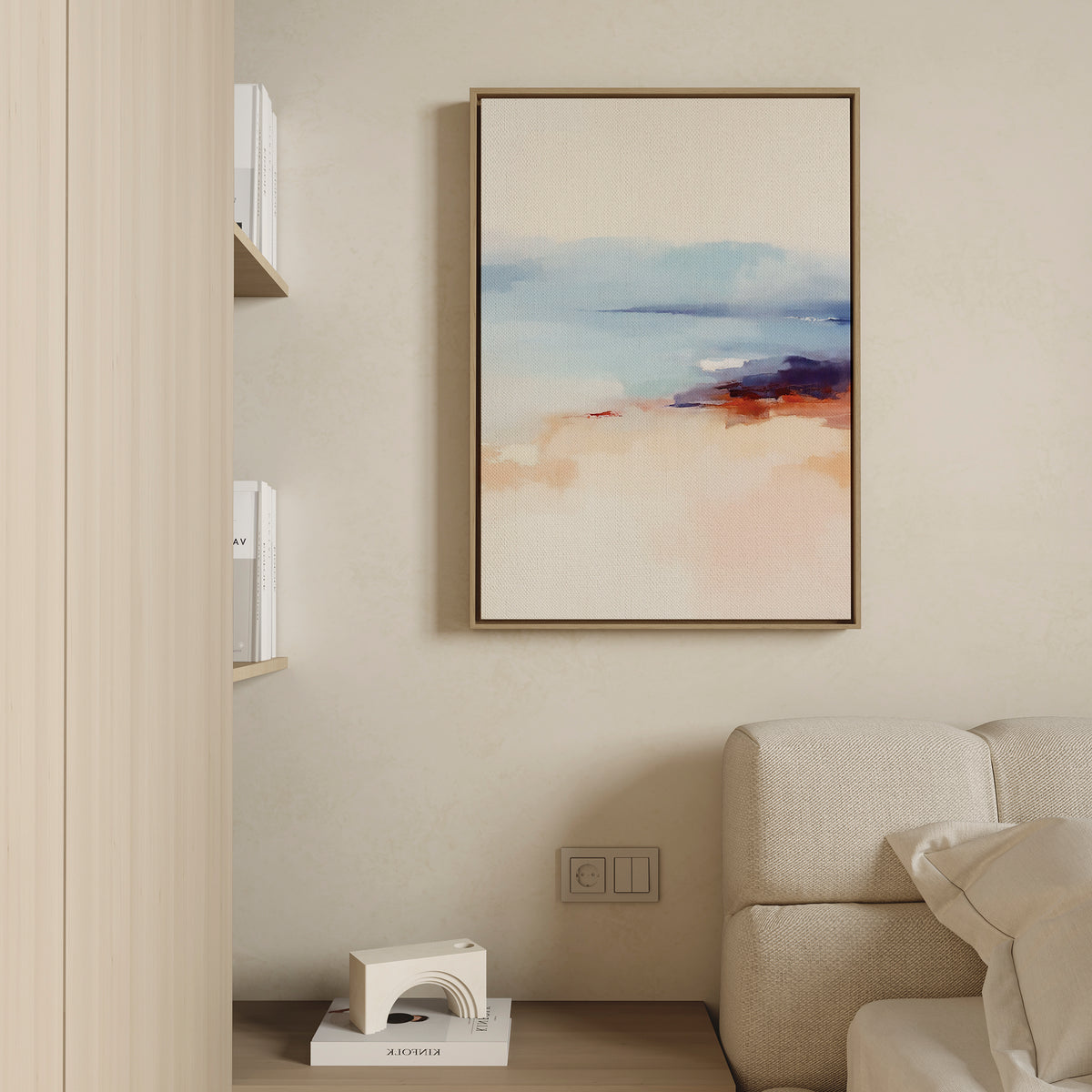 A minimalist living room features a beige wall, cream sofa, the Windswept Coastline Canvas Art - Vertical in soft blue, peach, and orange hues, a small shelf with books, and a decorative geometric object on a low table.