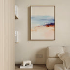 A minimalist living room features a beige wall, cream sofa, the Windswept Coastline Canvas Art - Vertical in soft blue, peach, and orange hues, a small shelf with books, and a decorative geometric object on a low table.
