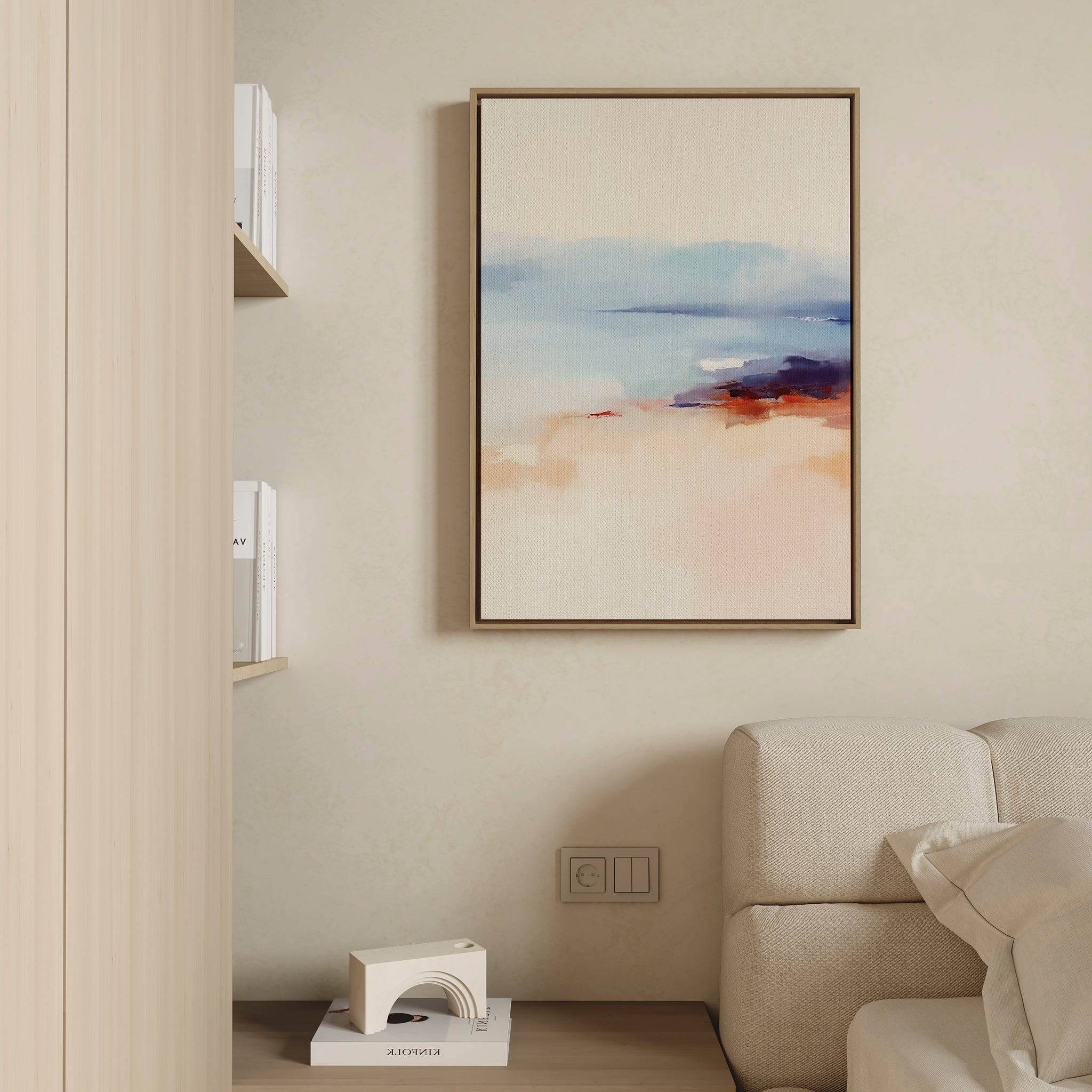 A minimalist living room features a beige wall, cream sofa, the Windswept Coastline Canvas Art - Vertical in soft blue, peach, and orange hues, a small shelf with books, and a decorative geometric object on a low table.