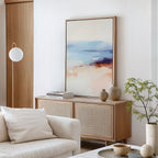 A modern living room with a light wood cabinet, the Windswept Coastline Canvas Art - Vertical as wall art, ceramic vases, books, a small plant, a round hanging lamp, and a white sofa with a cream pillow.