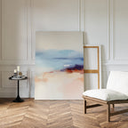 The Windswept Coastline Canvas Art - Vertical, featuring soft pastel blue, peach, and purple abstract tones, leans against a white paneled wall near a cushioned wooden chair and black side table on a herringbone wood floor.
