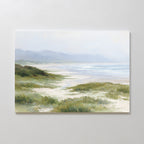 The Windswept Dunes Canvas Art - Horizontal features a tranquil coastal view with green dunes, coastal grasses, sandy beach, soft waves, and distant blue mountains beneath a pale sky—perfect for a calming space.