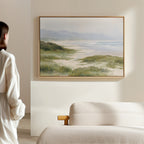 A person in white stands in a softly lit room, gazing at the Windswept Dunes Canvas Art - Horizontal featuring coastal grasslands and waves, displayed above a light beige sofa on a cream wall, creating a calming ambiance.