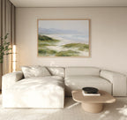 A modern living room with a beige sectional, wood coffee table, potted plant, and floor lamp features the "Windswept Dunes Canvas Art - Horizontal" above the sofa. Soft natural light enhances the calming coastal vibe.