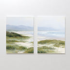 The "Windswept Dunes - Set of 2" features canvas paintings side by side, depicting a calm coastal landscape with sandy paths, grassy dunes, distant hills, and a tranquil ocean beneath a cloudy sky in soft, muted colors.