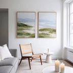 A modern living room with a gray sofa, woven chair, small white table with candles, and the Windswept Dunes - Set of 2 canvas prints creates a calming vibe as natural light pours in through large windows.