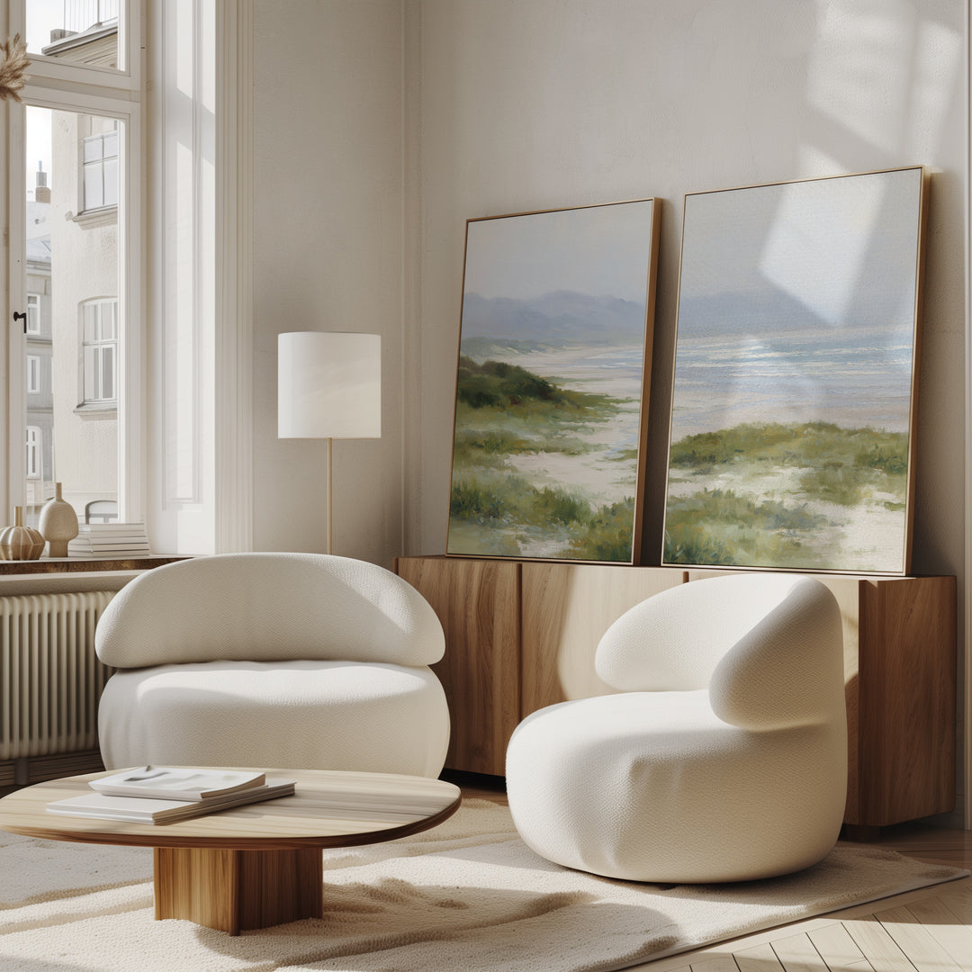 A modern living room with two rounded white armchairs, a low wooden coffee table, floor lamp, and the Windswept Dunes - Set of 2 canvas prints displayed on a wooden sideboard. Sunlight through large windows creates a calming atmosphere.