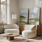 A modern living room with two rounded white armchairs, a low wooden coffee table, floor lamp, and the Windswept Dunes - Set of 2 canvas prints displayed on a wooden sideboard. Sunlight through large windows creates a calming atmosphere.