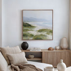A cozy living room features a beige sofa, neutral cushions, and a wooden sideboard with ceramic vases and books. The white wall above displays the Windswept Dunes Canvas Art - Square, adding to the calming atmosphere.