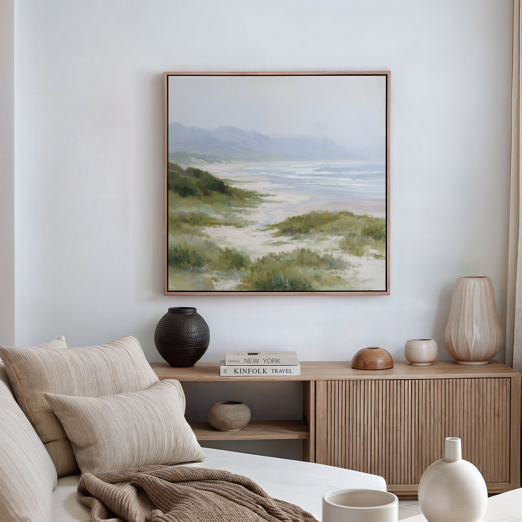 A cozy living room features a beige sofa, neutral cushions, and a wooden sideboard with ceramic vases and books. The white wall above displays the Windswept Dunes Canvas Art - Square, adding to the calming atmosphere.