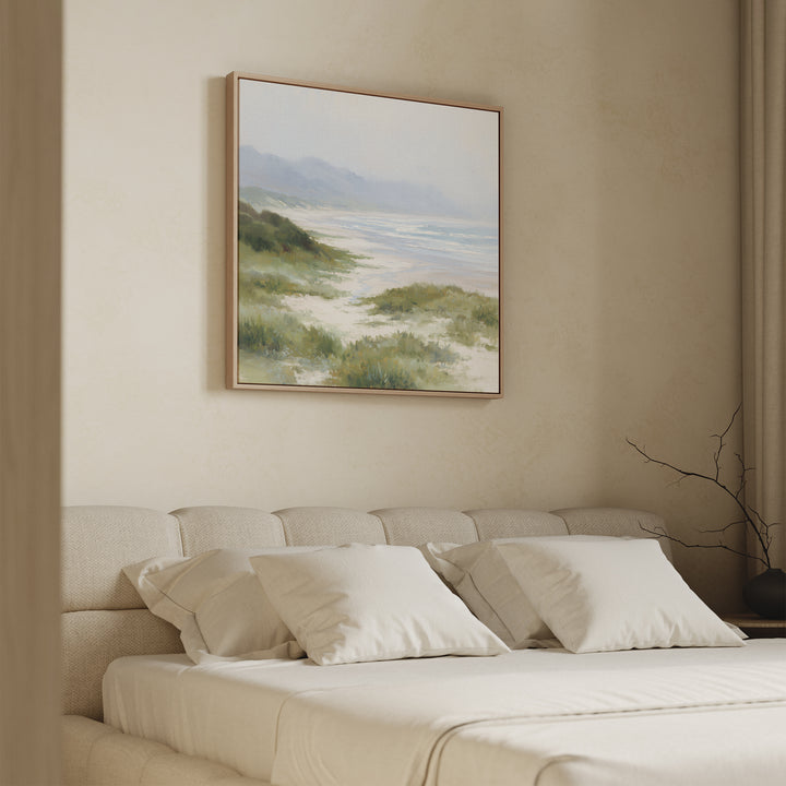 A beige bedroom features a cushioned headboard, cream bedding, and four pillows. Above the bed hangs the Windswept Dunes Canvas Art - Square, depicting tranquil coastal grasslands and waves. A bare branch rests on a small bedside table.