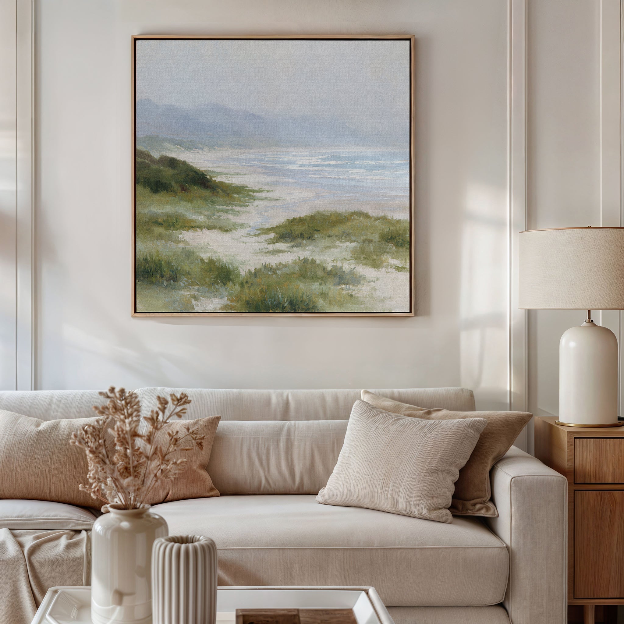 A cozy living room features a beige sofa, cream pillows, and a side table with a lamp. A vase of dried flowers adds charm, while the Windswept Dunes Canvas Art - Square brings a calming touch above the sofa.