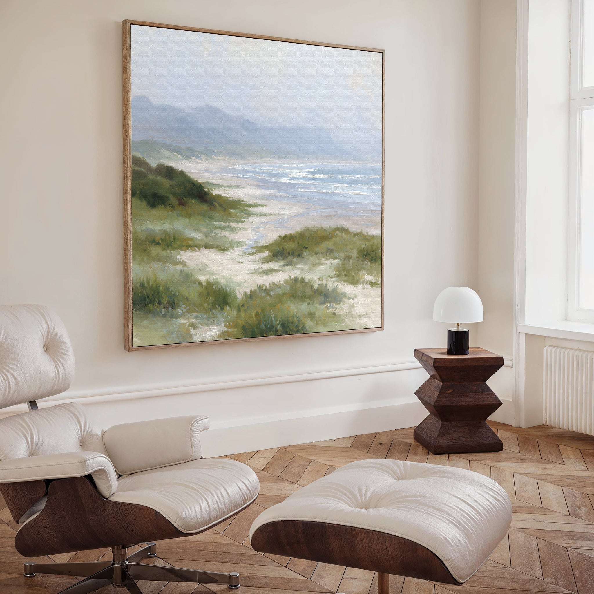 A cozy modern room with a white lounge chair, ottoman, wooden side table, and lamp features the Windswept Dunes Canvas Art - Square on a light wall, creating a calming atmosphere.