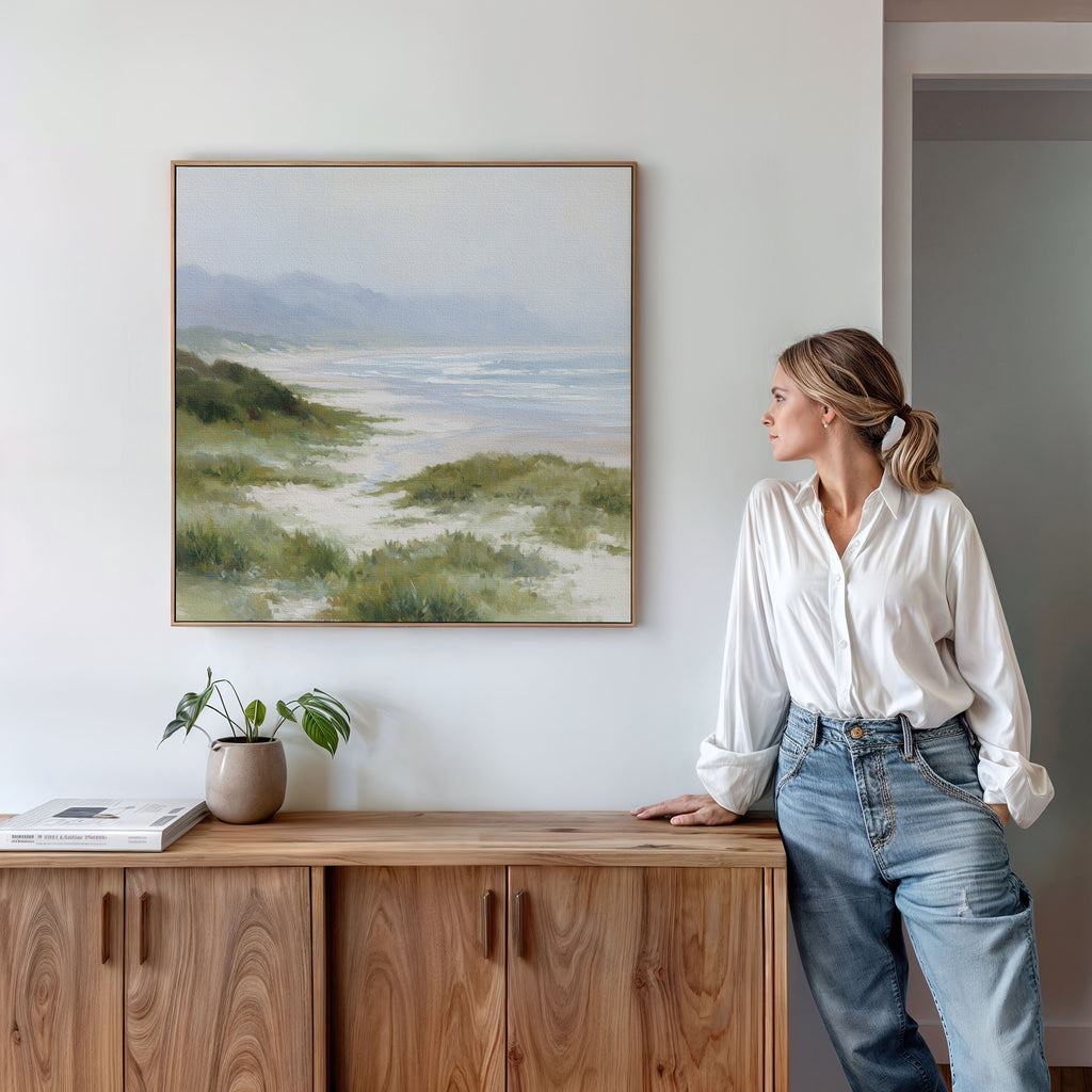 A woman in a white blouse and jeans leans against a wooden cabinet, gazing at the Windswept Dunes Canvas Art - Square displayed above her. A small potted plant and a book on the cabinet add to the serene ambiance.