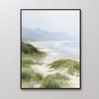 Windswept Dunes Canvas Art - Vertical features a serene beach scene with sandy shores, green grass, gentle waves, and misty mountains beneath a soft cloudy sky—perfect as calming coastal wall decor.