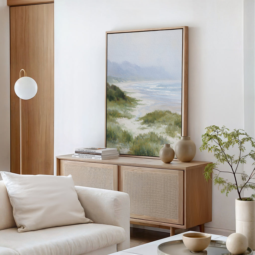 A minimal living room with a light wood sideboard, calming decor, neutral vases, a stack of books, a potted plant, and the Windswept Dunes Canvas Art - Vertical on a white wall. A cream sofa with a pillow sits in the foreground.
