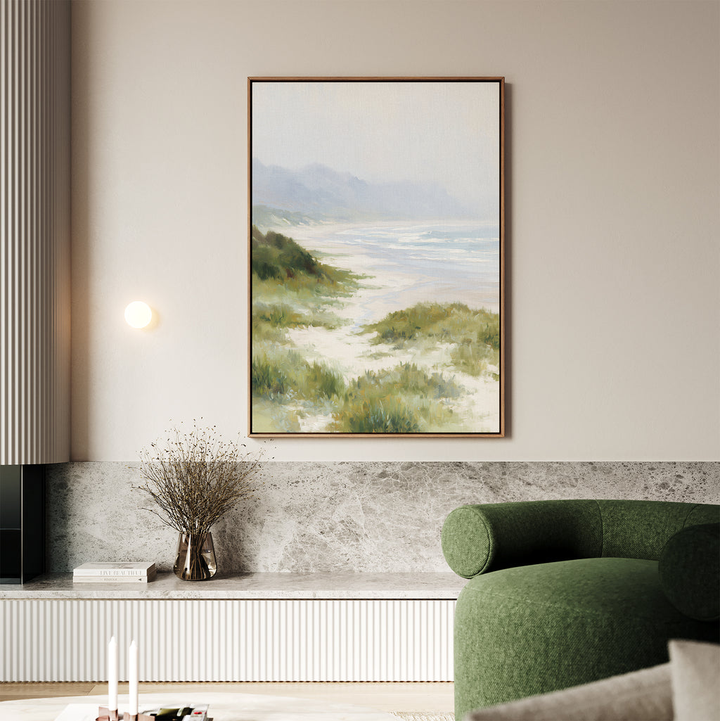 Add coastal tranquility to your space with the Windswept Dunes Canvas Art - Vertical, featuring sand dunes, grass, and distant mountains above a marble shelf in a modern living room with a green curved sofa and minimalist decor.