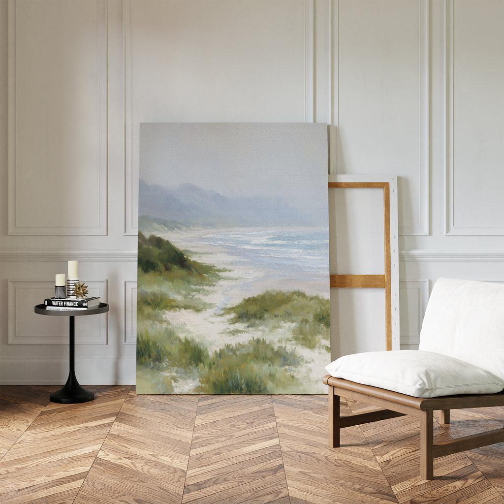 Windswept Dunes Canvas Art - Vertical leans against a white paneled wall in a minimalist room with wood floor, cushioned chair, and a black side table with candles and books—ideal for serene, coastal-inspired decor.