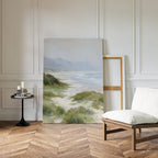 Windswept Dunes Canvas Art - Vertical leans against a white paneled wall in a minimalist room with wood floor, cushioned chair, and a black side table with candles and books—ideal for serene, coastal-inspired decor.