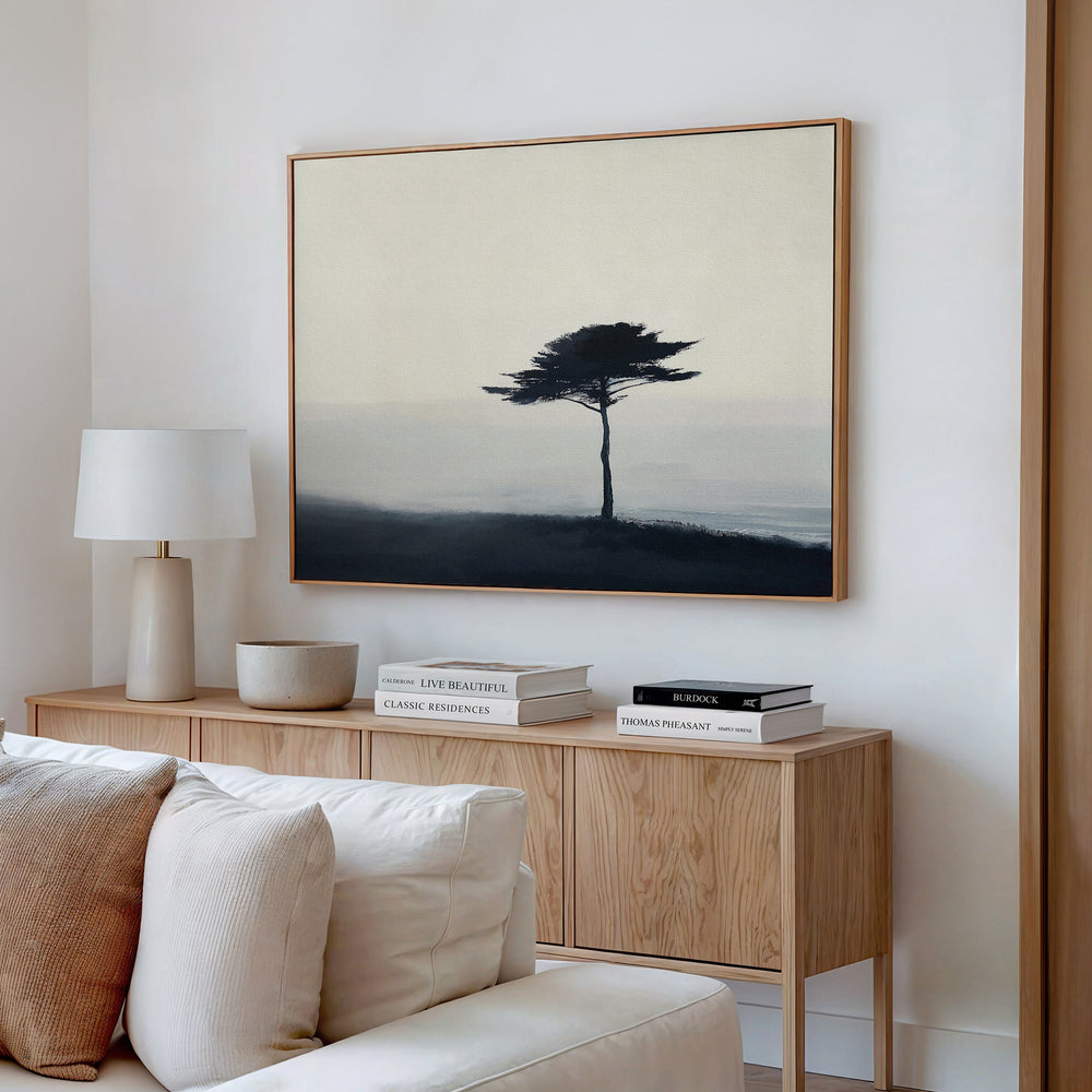 A minimalist living room showcases a light wood console with books and decor, a white table lamp, and the Windswept Solitude Canvas Art - Horizontal depicting a solitary tree in a misty landscape hanging above.