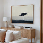 A minimalist living room showcases a light wood console with books and decor, a white table lamp, and the Windswept Solitude Canvas Art - Horizontal depicting a solitary tree in a misty landscape hanging above.
