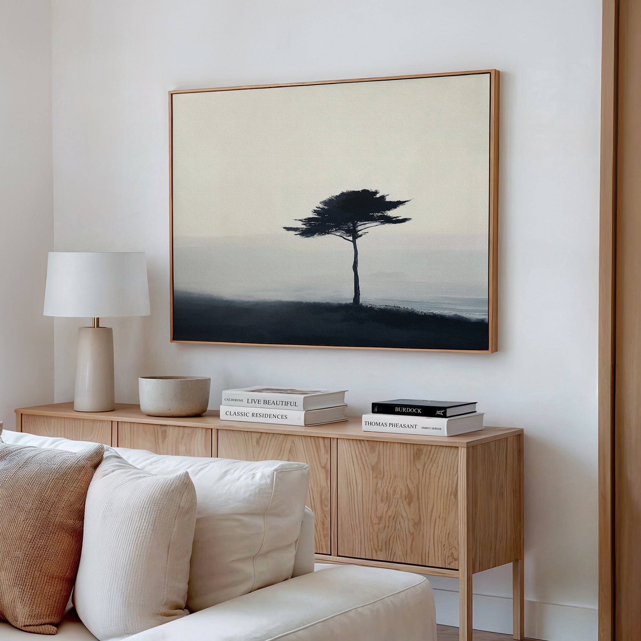 A minimalist living room showcases a light wood console with books and decor, a white table lamp, and the Windswept Solitude Canvas Art - Horizontal depicting a solitary tree in a misty landscape hanging above.