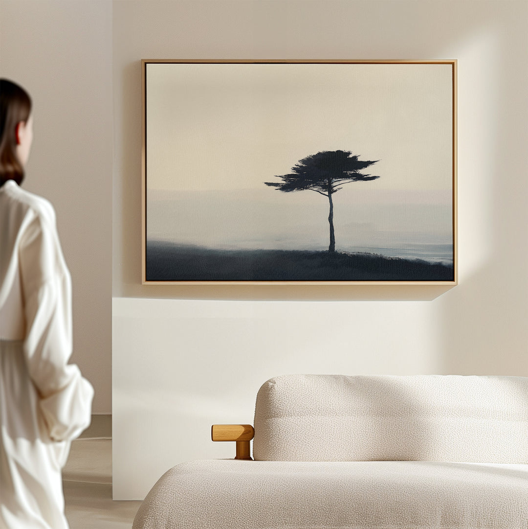 A person in a white robe stands in a minimal room with a light bed, admiring the Windswept Solitude Canvas Art - Horizontal, featuring a moody solitary tree—an ideal modern accent for living room decor.