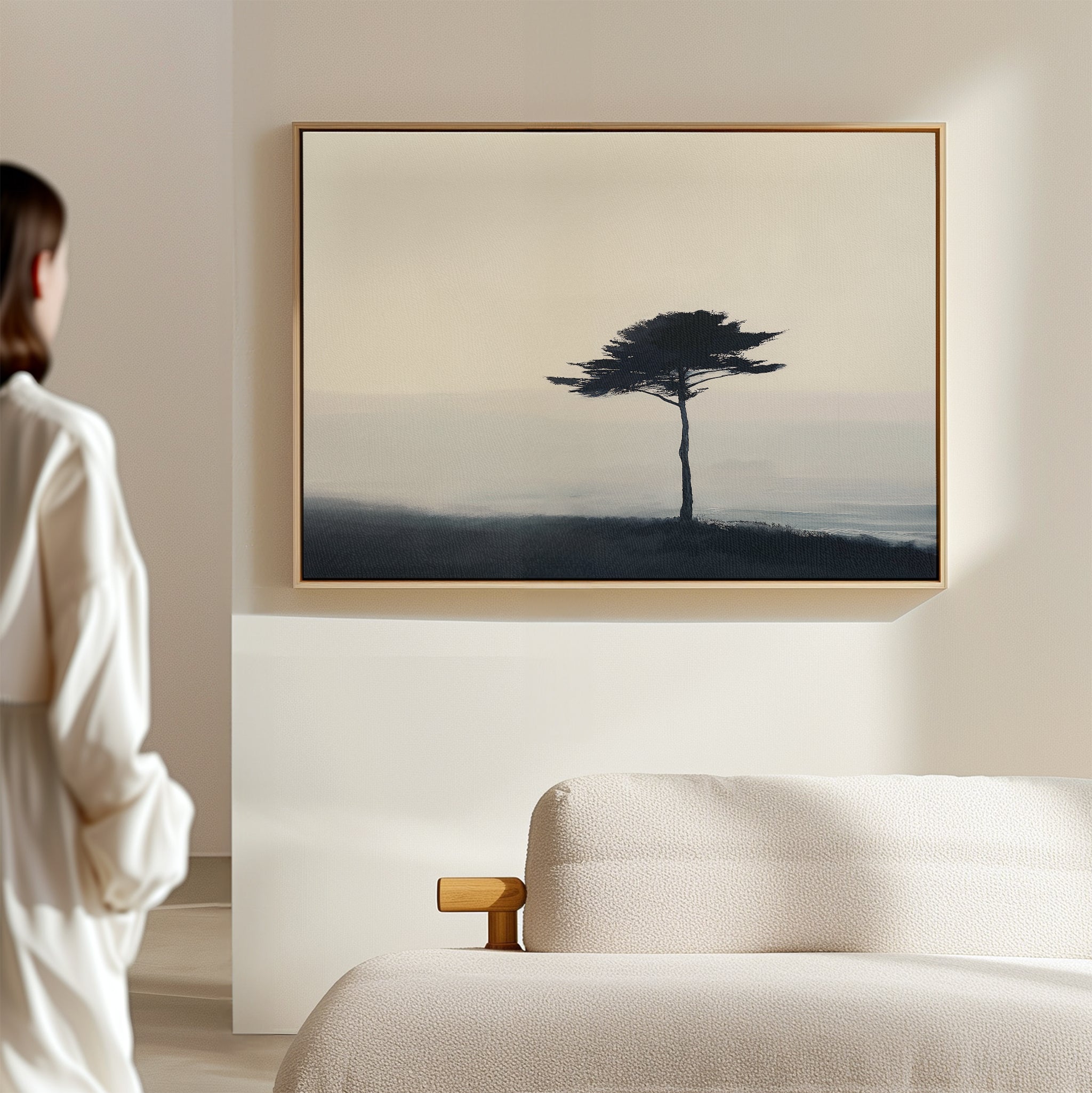 A person in a white robe stands in a minimal room with a light bed, admiring the Windswept Solitude Canvas Art - Horizontal, featuring a moody solitary tree—an ideal modern accent for living room decor.