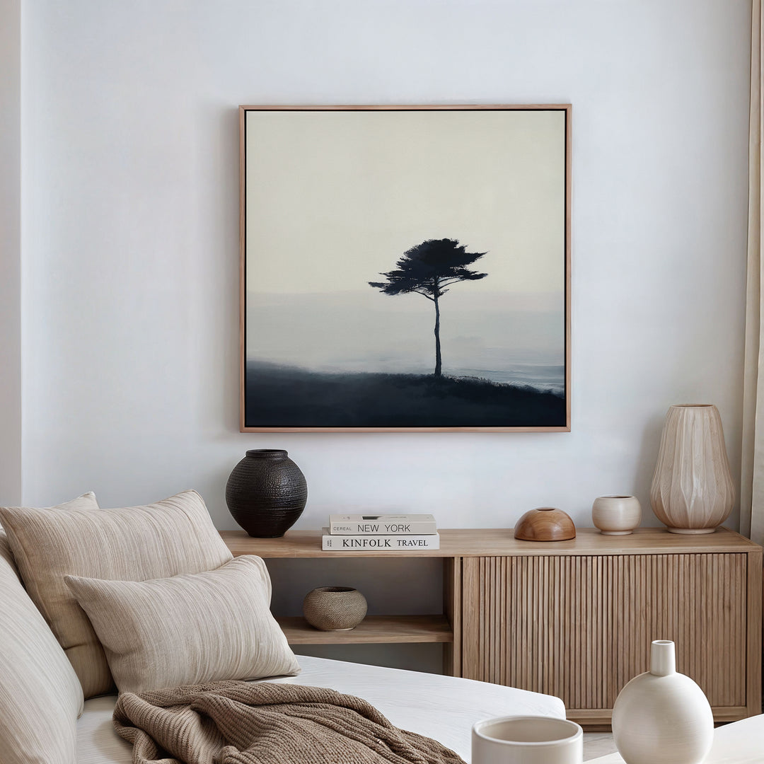 Minimalist living room with a light sofa, neutral pillows, wooden cabinet, ceramic vases, books, and the Windswept Solitude Canvas Art - Square—a striking moody print of a solitary tree in mist—perfect for modern decor.