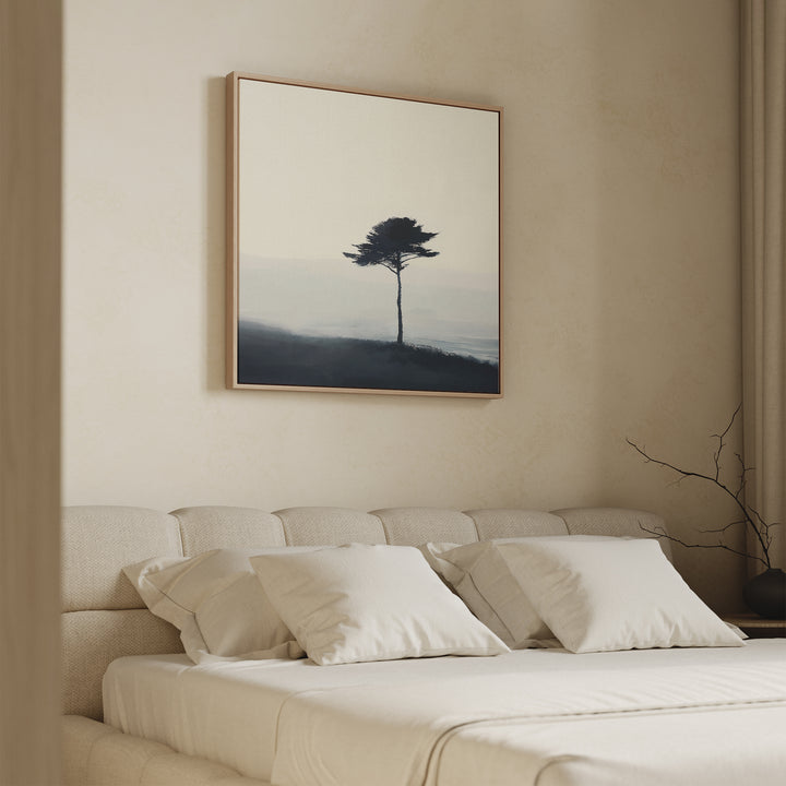 A minimalist bedroom with a beige upholstered bed, white bedding, and the Windswept Solitude Canvas Art - Square of a lone tree above the bed. A branch on the side table and neutral walls create a calm atmosphere.