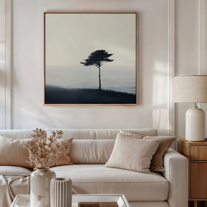 A neutral living room features a white sofa, beige pillows, a wooden side table with a ceramic lamp, and the Windswept Solitude Canvas Art - Square of a lone tree above the couch. Dried flowers in a vase enhance the modern decor.