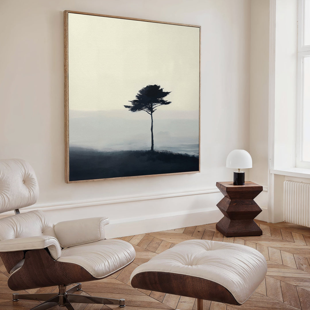 A modern, minimalist living room with a white lounge chair and ottoman, a geometric wooden side table with lamp, and the Windswept Solitude Canvas Art - Square as moody minimalist wall art featuring a solitary tree in misty landscape.