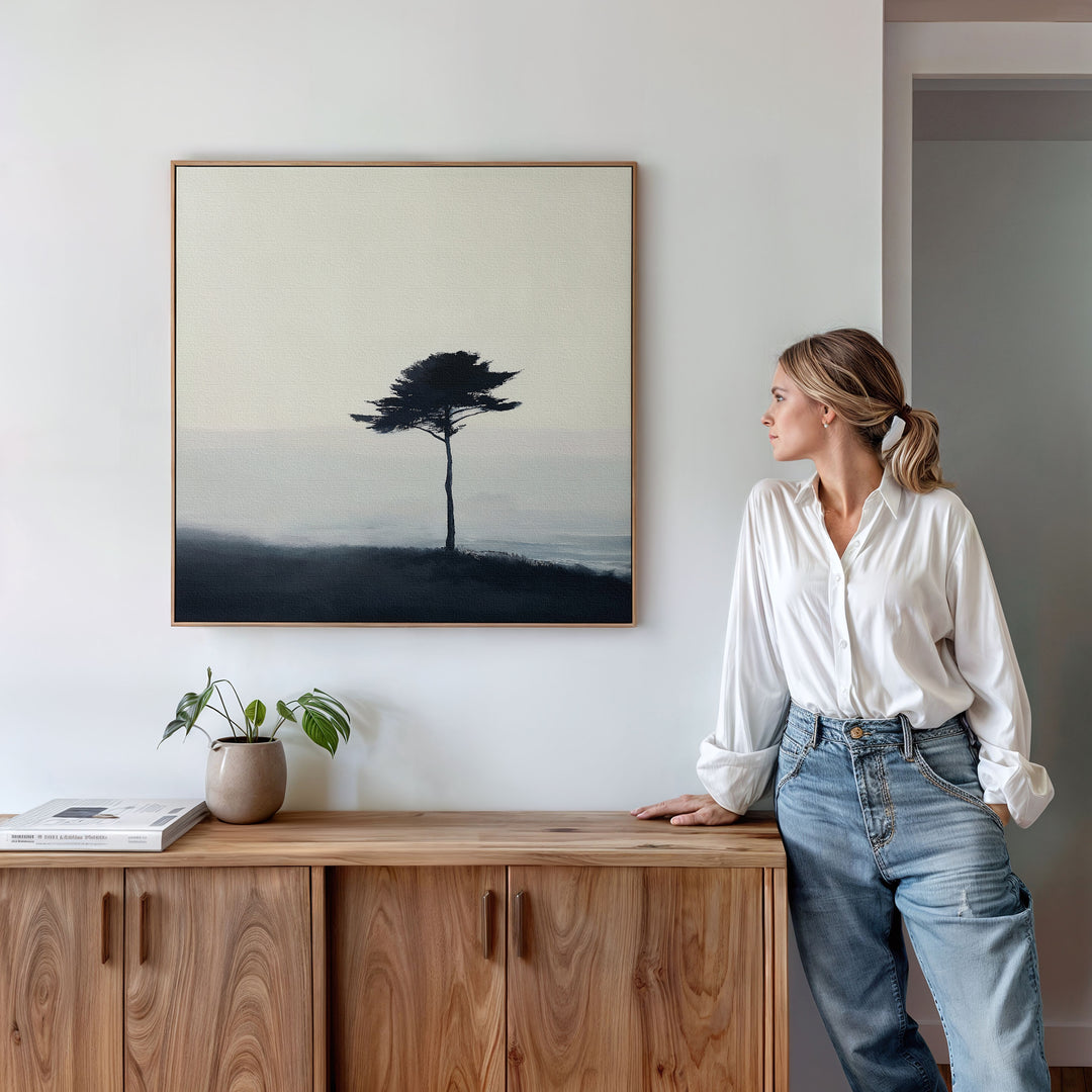 A woman in a white shirt and jeans stands by a wooden cabinet, admiring the Windswept Solitude Canvas Art - Square, which depicts a solitary tree in mist. A potted plant and book complete the modern living room decor.