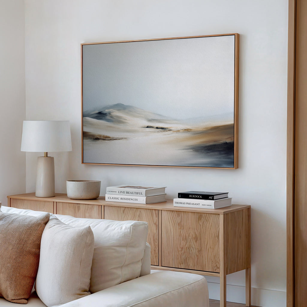A modern living room with a light wood sideboard, white table lamp, books, and bowl is styled with the horizontal Winter Plains Canvas Art in soft neutrals above. A light-colored sofa anchors the foreground.
