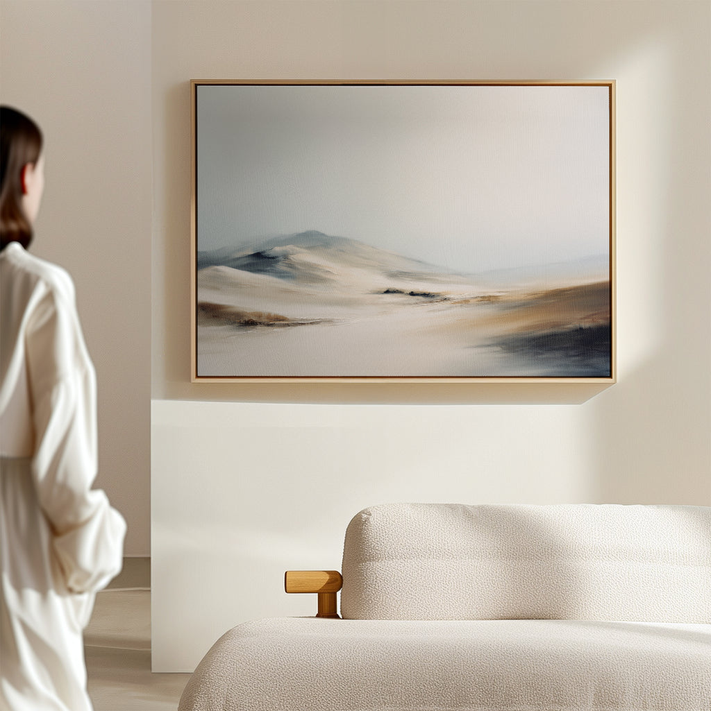 A person in a white shirt stands in a minimal, neutral-toned room, gazing at the Winter Plains Canvas Art - Horizontal, a framed abstract landscape with soft tones displayed above a textured beige couch.