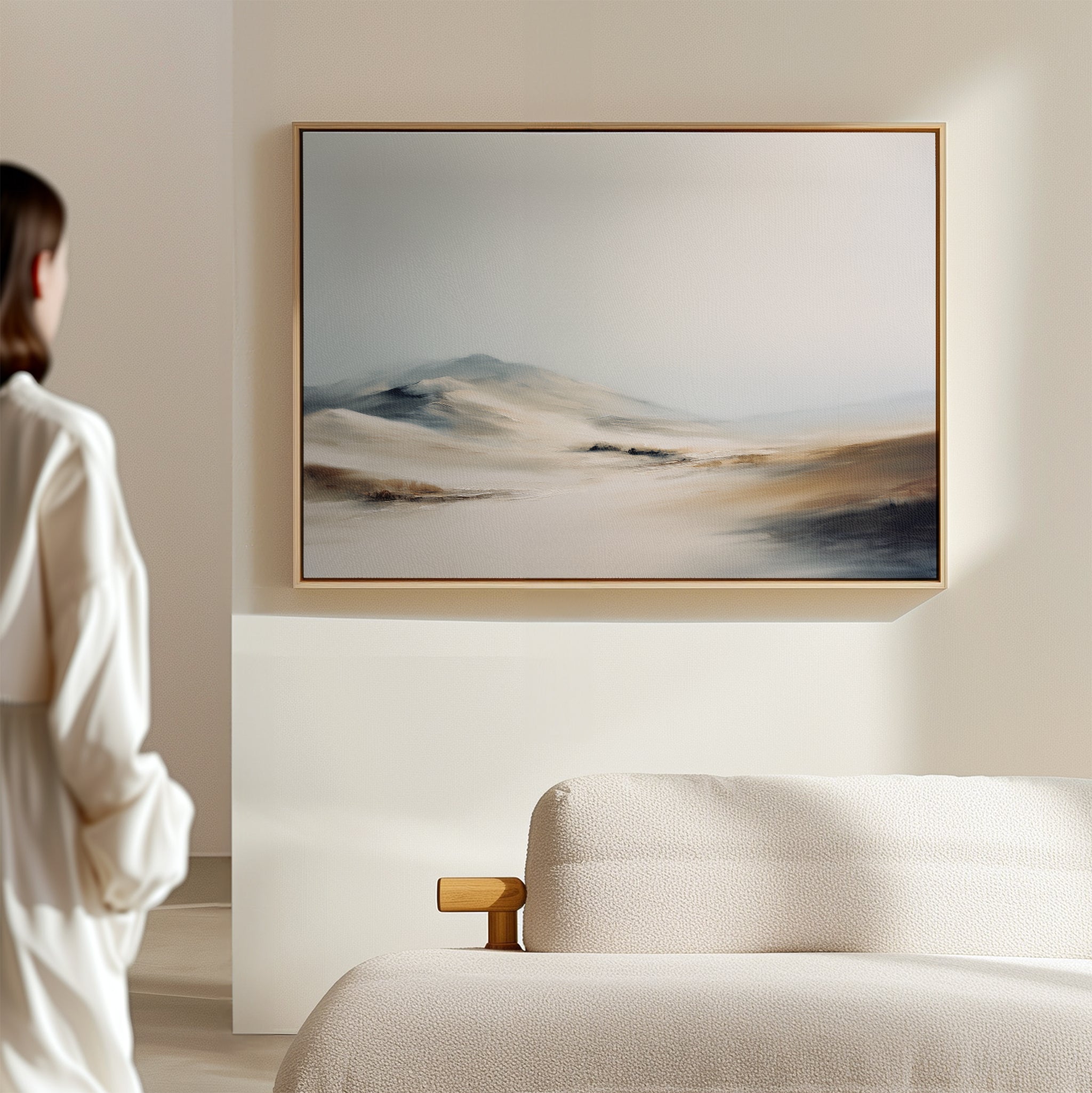 A person in a white shirt stands in a minimal, neutral-toned room, gazing at the Winter Plains Canvas Art - Horizontal, a framed abstract landscape with soft tones displayed above a textured beige couch.