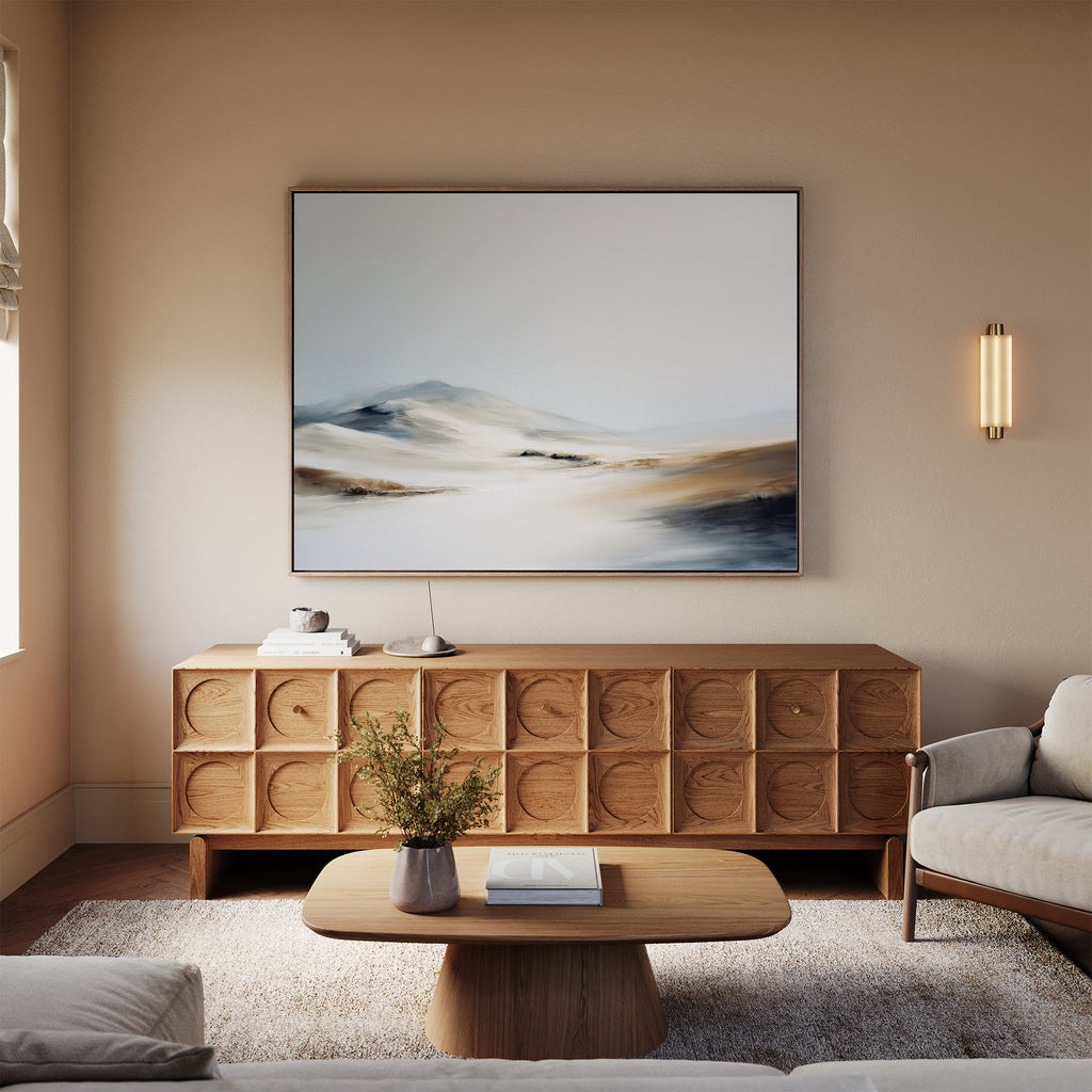 A modern living room features a wooden coffee table, textured sideboard, "Winter Plains Canvas Art - Horizontal" above neutral walls, a light sofa, and a small vase with greenery. Soft natural light fills the space.