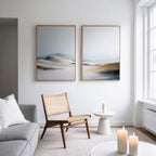 A modern, minimalist living room features a grey sofa, wooden chair, small white side table, and lit candles. Above the chair hang two "Winter Plains - Set of 2" wall art pieces. Natural light pours in from a large window.