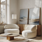 A modern living room with two white armchairs, a low wooden coffee table, and a wooden sideboard. The "Winter Plains - Set of 2" abstract landscape paintings rest against the wall as sunlight streams through large windows.