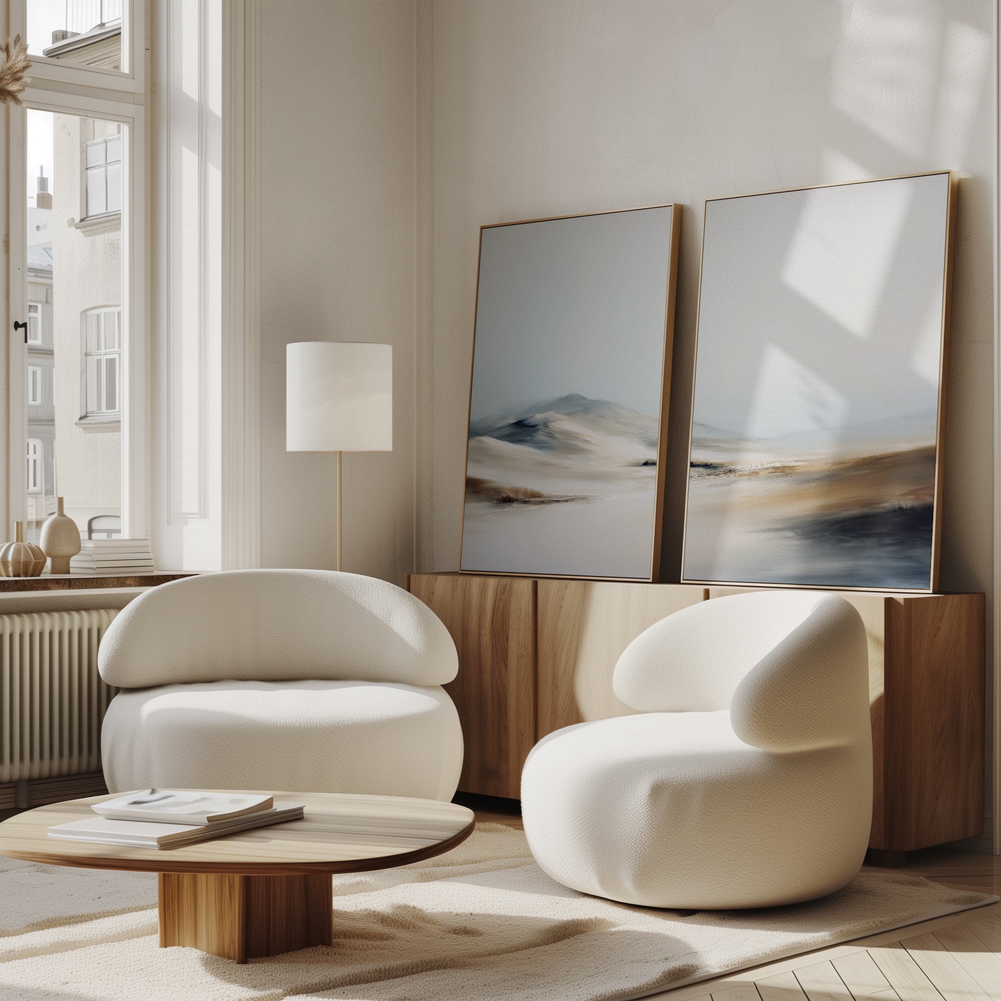 A modern living room with two white armchairs, a low wooden coffee table, and a wooden sideboard. The "Winter Plains - Set of 2" abstract landscape paintings rest against the wall as sunlight streams through large windows.