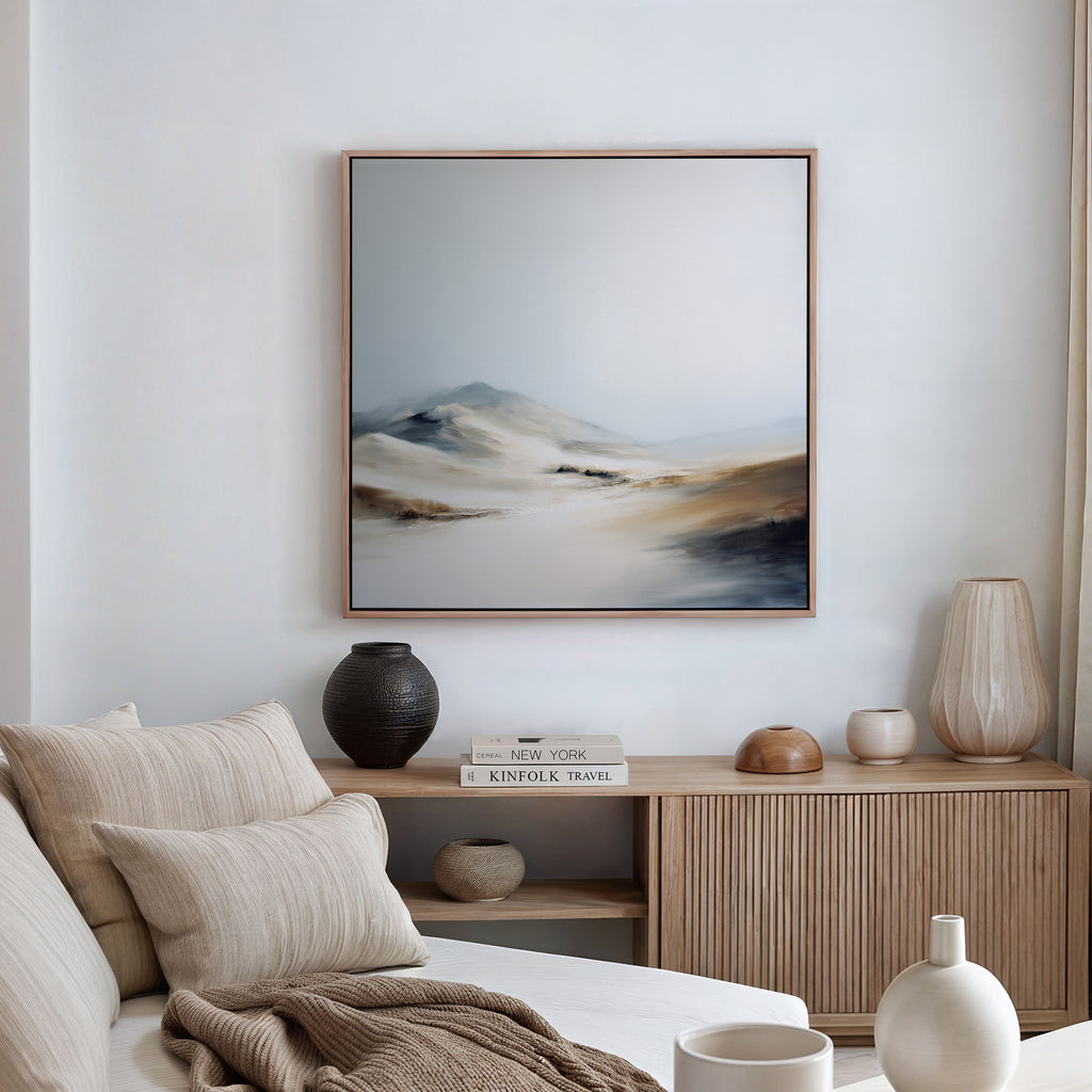 A minimalist living room with a beige sofa, neutral cushions, and a wooden sideboard. The Winter Plains Canvas Art - Square hangs above, while decorative vases and books on the sideboard complete the serene look.
