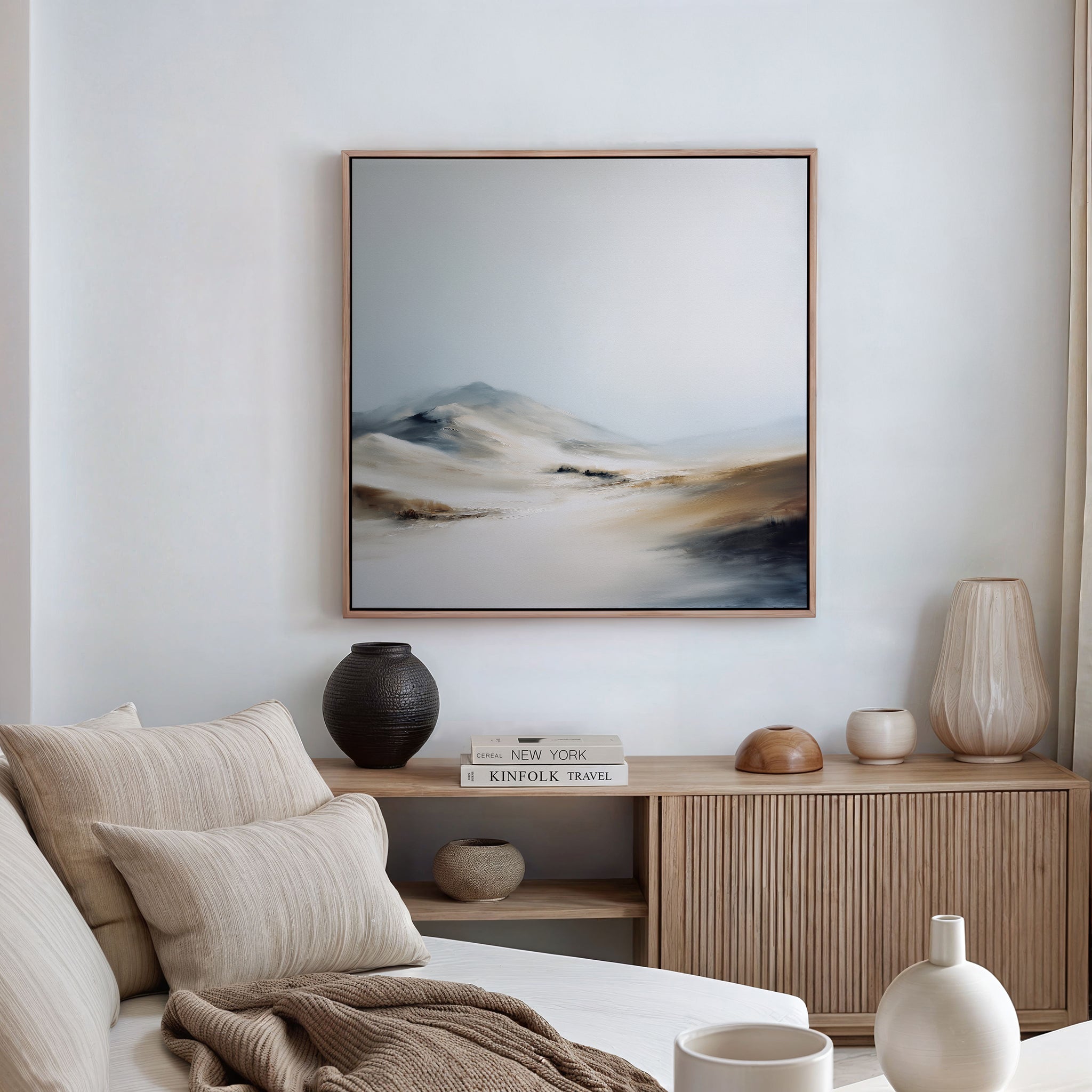 A minimalist living room with a beige sofa, neutral cushions, and a wooden sideboard. The Winter Plains Canvas Art - Square hangs above, while decorative vases and books on the sideboard complete the serene look.