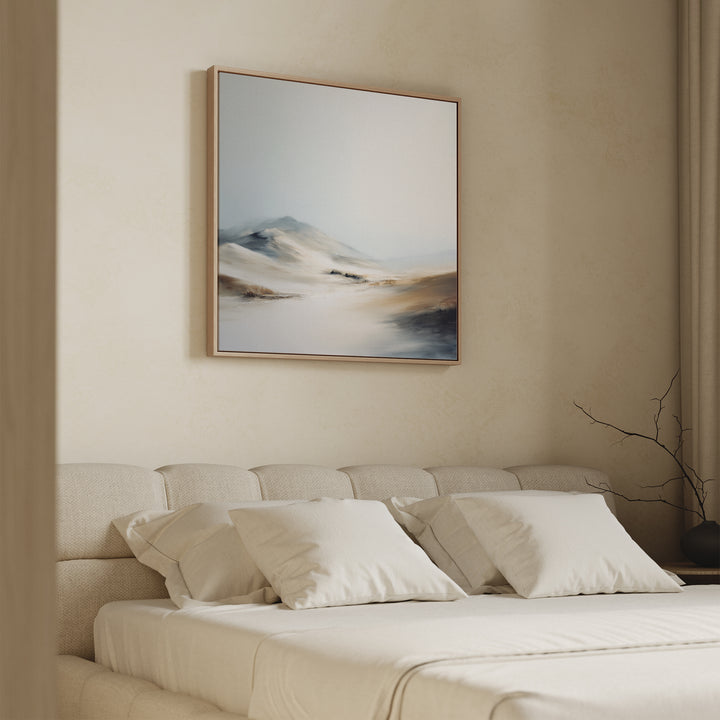 A softly lit bedroom with a beige bed, white pillows, and neutral bedding features the Winter Plains Canvas Art - Square above the bed—a serene abstract landscape in light blue, beige, and brown. A bare branch adorns the nightstand.