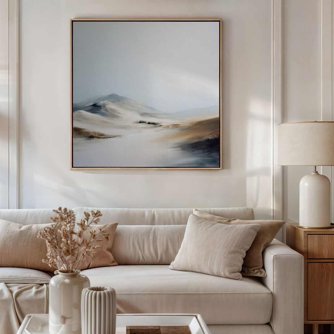 A modern living room with a beige sofa, neutral pillows, a side table and white lamp, dried flowers in a vase, and serene decor featuring the Winter Plains Canvas Art - Square framed on the wall.
