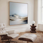 A modern, minimalist living room features a cream leather lounge chair and ottoman, wooden side table with white lamp, and the Winter Plains Canvas Art - Square above. Light wood herringbone flooring and a large window complete the airy space.