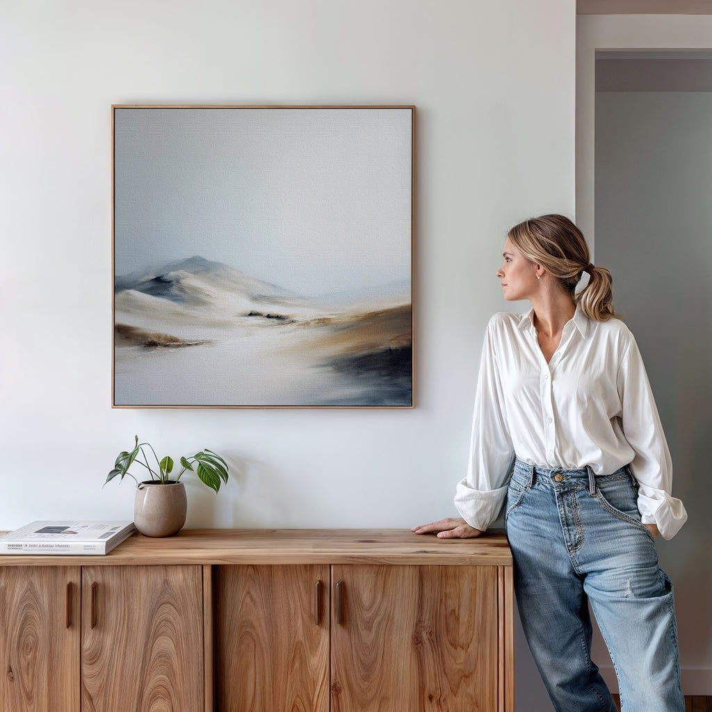 A woman in a white blouse and jeans stands by a wooden cabinet, admiring the Winter Plains Canvas Art - Square, a serene framed landscape painting of mountains and hills on a white wall. A potted plant and books sit atop the cabinet.