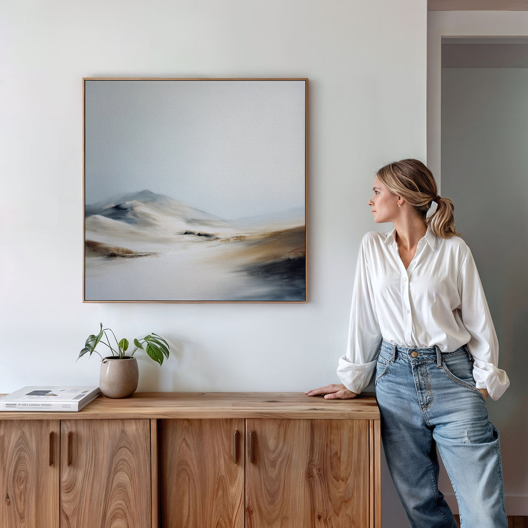 A woman in a white blouse and jeans stands by a wooden cabinet, admiring the Winter Plains Canvas Art - Square, a serene framed landscape painting of mountains and hills on a white wall. A potted plant and books sit atop the cabinet.