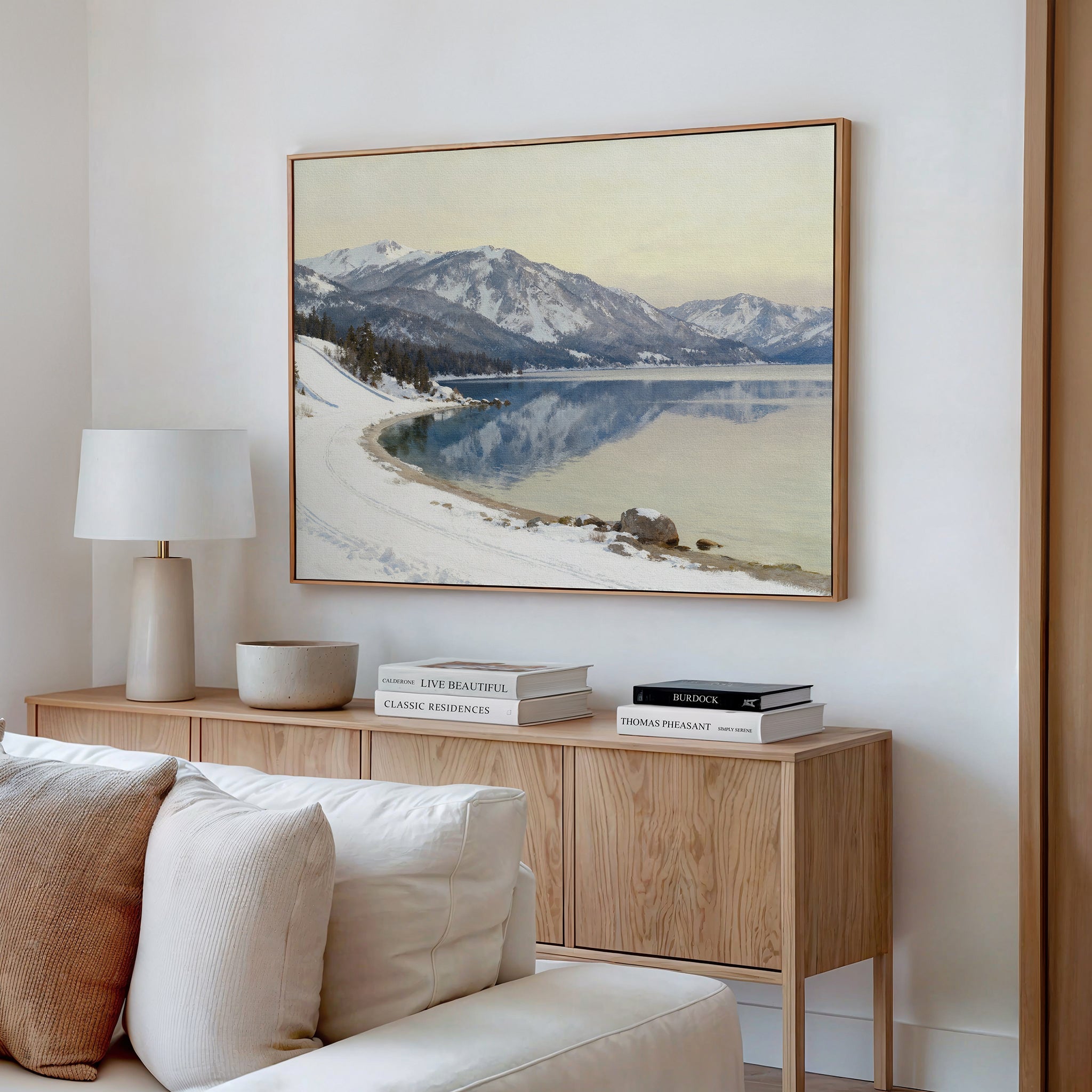 A cozy living room features a beige sofa, wooden sideboard with books and decor, white lamp, and serene landscape accents with the Winter Reflection Canvas Art - Horizontal showcasing a snowy mountain scene above an alpine lake.
